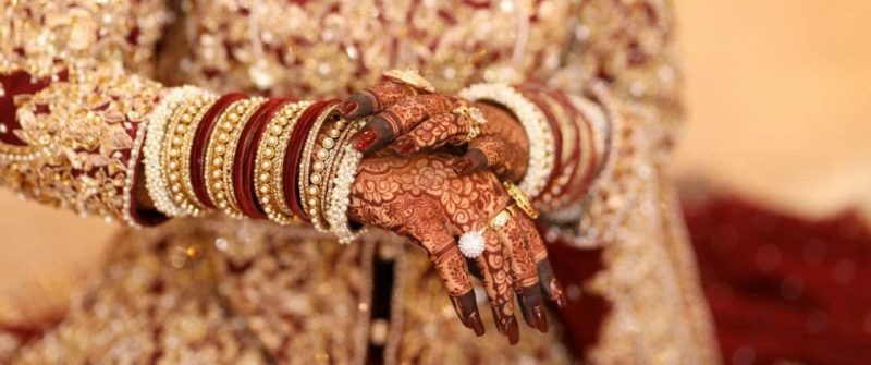Heavy full-hand bridal mehndi design with deep maroon stain and floral jaali patterns, paired with red and gold bridal bangles